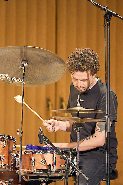 Ein Schlagzeuger mit lockigen Haaren und schwarzem T-Shirt spielt konzentriert an seinem Drumset in einem Tonstudio oder Aufnahmeraum. Das Schlagzeug besteht aus verschiedenen Becken und Trommeln, und im Hintergrund sind Holzwände und Mikrofonständer zu sehen.