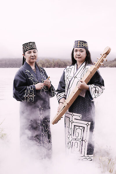 Das Bild zeigt zwei Frauen in traditioneller Ainu-Kleidung mit ornamentverzierten Gewändern und Kopfbedeckungen, die in einer nebligen Winterlandschaft stehen. Eine der Frauen hält ein traditionelles Saiteninstrument (Tonkori) in den Händen, während um sie herum mystischer Rauch oder Nebel aufsteigt.