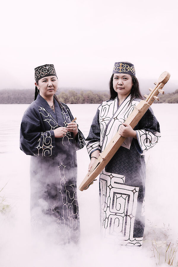The picture shows two women in traditional Ainu clothing with ornamental robes and headdresses, standing in a misty winter landscape. One of the women is holding a traditional stringed instrument (tonkori) in her hands, while mystical smoke or mist rises around her.
