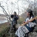 Das Bild zeigt zwei Frauen in traditioneller Ainu-Kleidung am Ufer eines Sees in einer herbstlichen Landschaft mit kahlen Bäumen. Eine Frau sitzt mit einem Tonkori-Saiteninstrument auf einem Stein, während die andere stehend in die Ferne blickt.