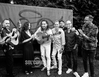 Das schwarz-weiße Foto zeigt eine Gruppe von sieben Musikern, die sich freundschaftlich umarmen und dabei Gitarren halten. Sie stehen vor einem Banner oder Plakat im Freien unter Bäumen und wirken fröhlich und entspannt nach einem gemeinsamen Auftritt oder Shooting.