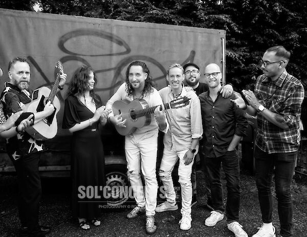 The black and white photo shows a group of seven musicians hugging each other amicably while holding guitars. They are standing in front of a banner or poster outdoors under trees and look cheerful and relaxed after a joint performance or shoot.