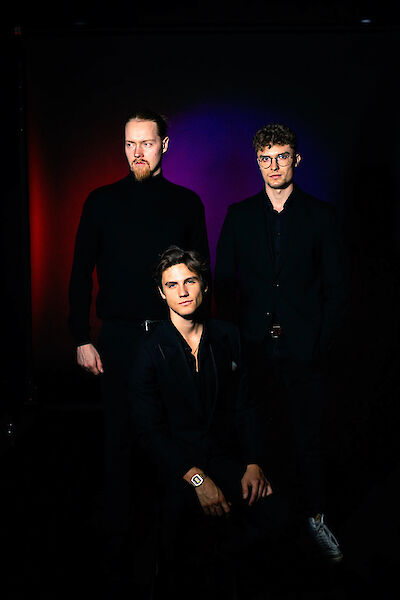 Das Bild zeigt drei junge Männer in schwarzer Kleidung, die vor einem dunklen Hintergrund mit violetter und roter Beleuchtung posieren. Ein Mann sitzt im Vordergrund, während zwei andere stehend hinter ihm arrangiert sind, was ein typisches Bandportrait darstellt.