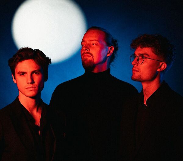 The image shows three men in black clothing posing in a professionally lit studio setting, with a large bright circle like a moon or spotlight in the background. The dramatic lighting with red and blue tones gives the band portrait an artistic, atmospheric mood.