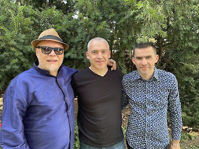 Das Bild zeigt drei Männer. Sie stehen zusammen. Sie lächeln in die Kamera. Der linke Mann trägt einen Hut. Er trägt auch eine Sonnenbrille. Der mittlere Mann trägt ein schwarzes T-Shirt. Der rechte Mann trägt ein Hemd mit Muster. Hinten sind grüne Bäume. Es sind Nadelbäume. Die Bäume sind der Hintergrund für das Foto.