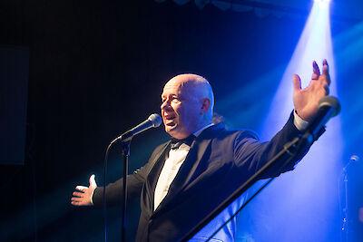 Ein Sänger trägt einen schwarzen Anzug. Er steht vor einem Mikrofon. Das Mikrofon ist auf einer Bühne. Der Sänger hebt eine Hand hoch. Er zeigt viel Gefühl. Blaues Licht leuchtet auf der Bühne. Das Licht macht eine besondere Stimmung. Es ist ein Live-Konzert. Die Atmosphäre ist sehr stark.
