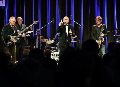 Eine Band spielt auf einer Bühne. Die Bühne hat blaues Licht. Vier Musiker sind da. Der Sänger steht in der Mitte. Er trägt einen schwarzen Anzug. Die anderen Musiker spielen Gitarre und Saxophon. Viele Menschen schauen zu. Man sieht nur ihre Schatten. Hinten steht ein Schlagzeug. Auf dem Schlagzeug steht "Jazz Schmiede".