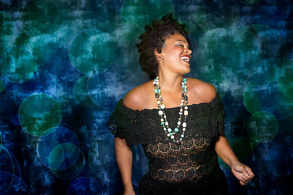 A cheerful woman with short curls wears a black off-the-shoulder dress and an eye-catching multi-strand pearl necklace as she laughs and looks to the side. The background shows an artistic bluish-green bokeh effect with blurred circles of light, creating a festive and elegant atmosphere.