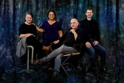 Vier Personen sitzen auf Stühlen. Der Hintergrund ist blau. Sie lächeln in die Kamera. Es sind zwei Frauen und zwei Männer. Sie tragen dunkle Kleidung. Sie wirken entspannt und professionell.