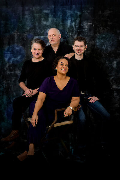 Vier Personen stehen zusammen für ein Foto. Eine Frau trägt ein violettes Shirt. Sie sitzt auf einem Stuhl. Drei Männer stehen hinter ihr. Sie tragen schwarze Kleidung. Der Hintergrund ist dunkel. Alle Personen lächeln. Das Foto sieht professionell aus. Die Stimmung ist freundlich.