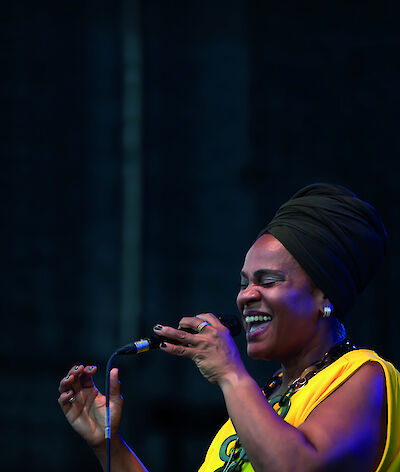 Eine Sängerin mit einem dunklen Kopftuch und einem gelben Oberteil steht mit einem Mikrofon auf der Bühne und singt mit geschlossenen Augen und einem Lächeln. Die Bühnenszenerie ist in atmosphärisches blaues Licht getaucht, das eine intime Konzertatmosphäre schafft.