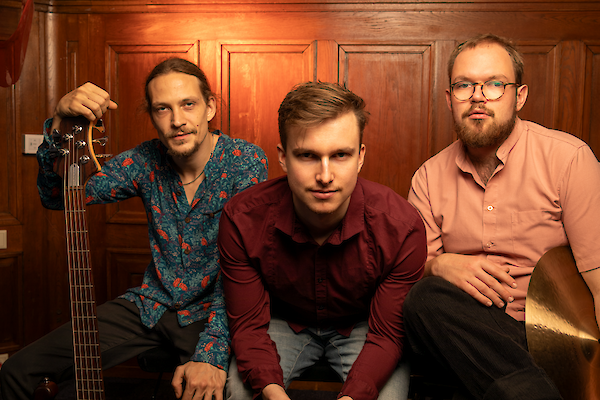 The picture shows three young men posing as a music band in front of a wood-panelled wall, with the man on the left holding a guitar. The musicians are wearing different coloured shirts - a patterned blue shirt, a dark red shirt and a pink shirt - and are looking at the camera in a friendly manner.