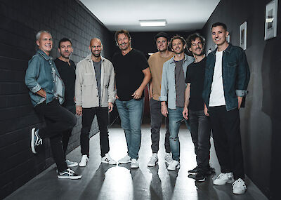 Acht Männer verschiedenen Alters stehen lächelnd nebeneinander in einem modernen Flur mit dunklen Wänden und hellem Boden. Die Gruppe ist lässig in Jeans, T-Shirts und Hemden gekleidet und posiert entspannt für ein Bandfoto in dem industriell anmutenden Innenraum.