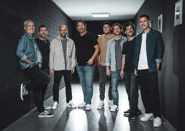 Acht Männer verschiedenen Alters stehen lächelnd nebeneinander in einem modernen Flur mit dunklen Wänden und hellem Boden. Die Gruppe ist lässig in Jeans, T-Shirts und Hemden gekleidet und posiert entspannt für ein Bandfoto in dem industriell anmutenden Innenraum.