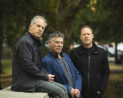 Das Bild zeigt drei Männer mittleren Alters, die nebeneinander in einem Park oder einer ähnlichen Außenumgebung posieren. Der linke Mann trägt eine schwarze Jacke, der mittlere einen blauen Anzug mit gemustertem Hemd und der rechte Mann eine schwarze Jacke, während im Hintergrund grüne Bäume und unscharf abgebildete Fahrzeuge zu sehen sind.