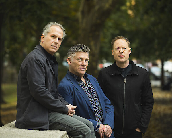 Das Bild zeigt drei Männer mittleren Alters, die nebeneinander in einem Park oder einer ähnlichen Außenumgebung posieren. Der linke Mann trägt eine schwarze Jacke, der mittlere einen blauen Anzug mit gemustertem Hemd und der rechte Mann eine schwarze Jacke, während im Hintergrund grüne Bäume und unscharf abgebildete Fahrzeuge zu sehen sind.