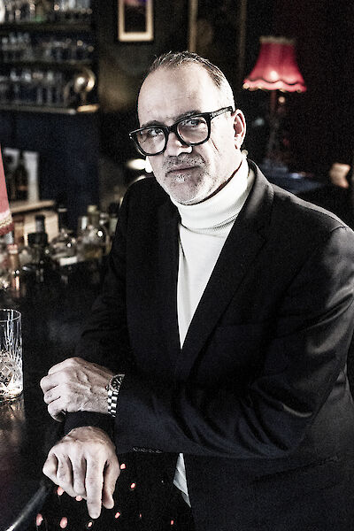 Das Bild zeigt einen eleganten Mann mittleren Alters mit Brille, der einen schwarzen Anzug über einem weißen Rollkragenpullover trägt und lächelnd in die Kamera blickt. Er sitzt in einer stilvoll eingerichteten Bar oder Lounge mit warmem Licht, einem Kristallglas vor sich und einer roten Lampe im Hintergrund.