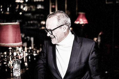 Das Bild zeigt einen Mann mit Brille und weißem Rollkragenpullover unter einem dunklen Anzug, der lächelnd zur Seite blickt. Er befindet sich in einer stimmungsvoll beleuchteten Bar-Atmosphäre mit roten Lampenschirmen und Kristallgläsern im Hintergrund.