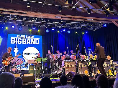 Das Bild zeigt eine Big Band. Die Band heißt Klaus Esser Big Band. Sie spielen auf einer Bühne. Die Bühne hat blaues Licht. Im Hintergrund hängt ein Banner. Auf dem Banner steht der Name der Band. Die Musiker sitzen auf der Bühne. Sie haben ihre Instrumente dabei. Vor jedem Musiker steht ein Notenständer. Auf den Ständern ist das Logo der Band. Ein Dirigent leitet die Band. Im Vordergrund sieht man das Publikum.