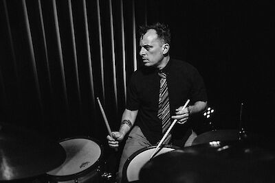 Das Schwarz-Weiß-Foto zeigt einen Schlagzeuger während einer Live-Performance, der konzentriert mit Drumsticks in beiden Händen an seinem Schlagzeug spielt. Im Hintergrund sind vertikale Strukturelemente oder Vorhänge zu erkennen, die eine Bühnenatmosphäre schaffen.