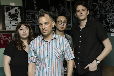 Das Bild zeigt vier Personen, die als Musikband für ein Gruppenfoto vor einer Wand mit verschiedenen Postern und Grafiken posieren. Die Gruppe besteht aus einer Frau und drei Männern unterschiedlichen Alters, die leger gekleidet sind und direkt in die Kamera blicken.