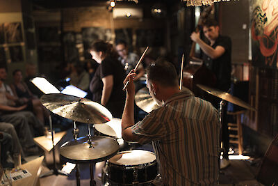 Das Bild zeigt einen Schlagzeuger in einem gestreiften Hemd, der während einer Live-Performance oder Probe in einem intimen Veranstaltungsraum spielt. Im Hintergrund sind weitere Musiker und Zuschauer zu erkennen, die in dem warmbeleuchteten Raum mit Postern an den Wänden eine entspannte Atmosphäre schaffen.