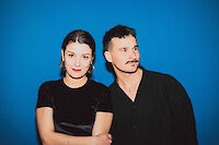 The picture shows two people against a bright blue background - a woman in a black T-shirt with her hair tied back and a man with a moustache in a black shirt. Both are looking directly at the camera and standing close together with their arms crossed.