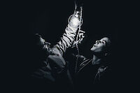 The black and white photo shows a person holding up a bottle and looking upwards, while a second person can be seen in the background. The dramatic lighting and dark background give the scene a theatrical, artistic atmosphere.