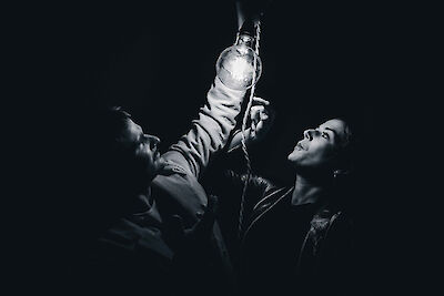 Das Schwarz-Weiß-Foto zeigt eine Person, die eine Flasche in die Höhe hält und dabei nach oben blickt, während eine zweite Person im Hintergrund zu sehen ist. Die dramatische Beleuchtung und der dunkle Hintergrund verleihen der Szene eine theatralische, künstlerische Atmosphäre.