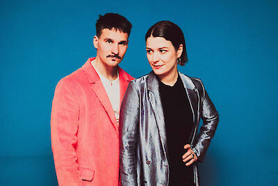 Das Bild zeigt zwei Personen vor einem leuchtend blauen Hintergrund - ein Mann mit Schnurrbart in einem rosa/korallenfarbenen Anzug und eine Frau in einer silbernen Lederjacke über schwarzem Oberteil. Beide posieren selbstbewusst für die Kamera und schauen direkt in das Objektiv.