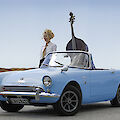 A blonde woman stands next to a light blue vintage convertible and holds a large double bass sticking out of the car. The scene takes place on a country road in front of a wide field under a blue sky.