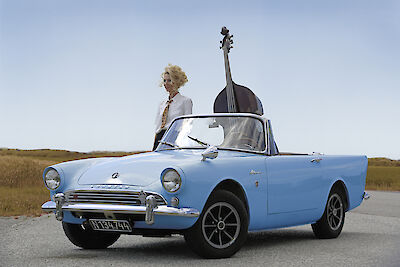 Eine blonde Frau steht neben einem hellblauen Oldtimer-Cabrio und hält einen großen Kontrabass, der aus dem Auto ragt. Die Szene spielt sich auf einer Landstraße vor einem weiten Feld unter blauem Himmel ab.