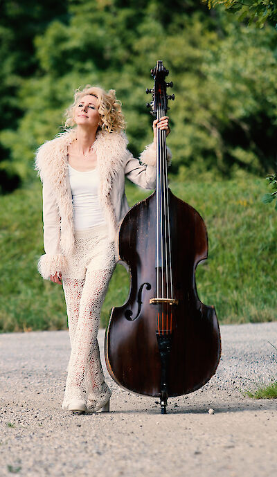 Eine blonde Frau mit lockigen Haaren steht neben einem großen Kontrabass und trägt ein elegantes weißes Outfit mit einer flauschigen beigen Weste. Die Aufnahme zeigt sie in einer natürlichen Parklandschaft mit grünen Bäumen im Hintergrund auf einem gepflasterten Weg.