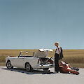 A blonde woman stands next to a white vintage convertible with the boot open on a country road. Next to the car are several cases of musical instruments, including a large double bass case, against the backdrop of a wide field landscape under a blue sky.