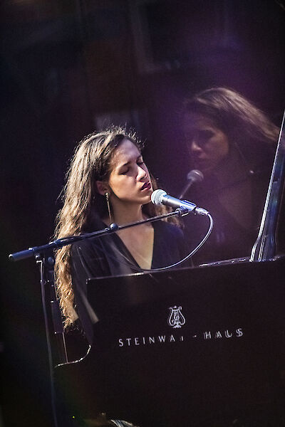 The picture shows a young woman with long brunette hair sitting at a black Steinway grand piano and singing into a microphone. The scene is atmospherically lit with purple stage lighting, suggesting a live performance or concert.