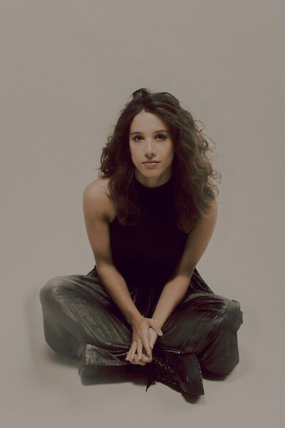 The picture shows a young woman with curly brown hair sitting cross-legged on the floor and looking directly into the camera. She is wearing a black sleeveless top and olive green loose trousers while posing in front of a neutral beige background.