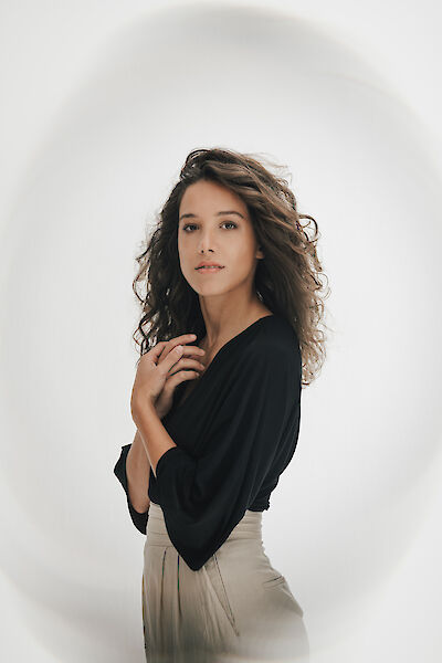 The picture shows a young woman with curly brunette hair wearing a black top and beige trousers. She poses in front of a neutral white background and looks directly into the camera with one hand on her hair.