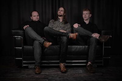 Drei junge Männer sitzen nebeneinander auf einem schwarzen Ledersofa vor einem dunklen Hintergrund und posieren für ein professionelles Foto. Sie tragen stilvolle, dunkle Kleidung und wirken selbstbewusst, was auf ein Bandfoto oder ein professionelles Gruppenporträt hindeutet.