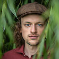 Das Bild zeigt das Porträt eines jungen Mannes mit langen Haaren und einer braunen Schiebermütze, der vor einem unscharfen grünen Pflanzenhintergrund steht. Er trägt ein rotes Hemd und blickt direkt in die Kamera, während grüne Blätter im Vordergrund das Bild rahmen.