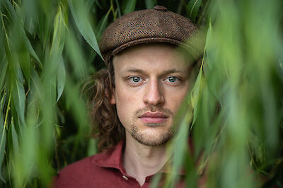 Das Bild zeigt das Porträt eines jungen Mannes mit langen Haaren und einer braunen Schiebermütze, der vor einem unscharfen grünen Pflanzenhintergrund steht. Er trägt ein rotes Hemd und blickt direkt in die Kamera, während grüne Blätter im Vordergrund das Bild rahmen.