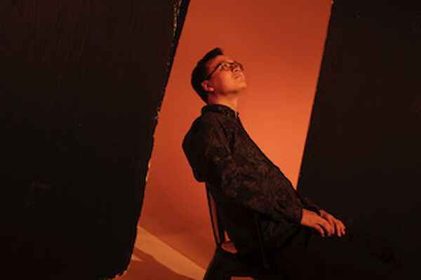 Ein junger Mann mit Brille und dunkler Jacke posiert zwischen zwei schwarzen Vorhängen vor einem warm-orangefarbenen Hintergrund. Er blickt nach oben und lehnt sich leicht zurück, was dem Bild eine dramatische und stimmungsvolle Atmosphäre verleiht.