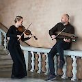 The picture shows two musicians in a historic building with a stone balustrade: a woman dressed in black plays the violin while a man in a dark jumper holds a nyckelharpa. The two musicians are in an elegant architectural setting with cream-coloured walls and white balustrades, reminiscent of a castle or historic building.