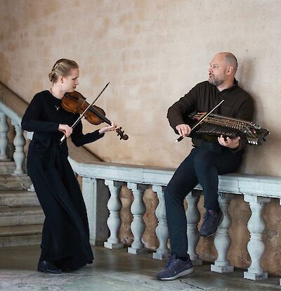 Das Bild zeigt zwei Musiker in einem historischen Gebäude mit Steinbalustrade: eine Frau in schwarzer Kleidung spielt Violine, während ein Mann in dunklem Pullover eine Nyckelharpa hält. Die beiden Musiker befinden sich in einer eleganten Architektur mit cremefarbenen Wänden und weißen Balustraden, die an ein Schloss oder historisches Gebäude erinnert.