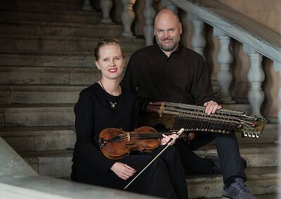 Das Bild zeigt ein Musikerduo bestehend aus einer Frau in schwarzer Kleidung mit einer Violine und einem Mann mit Vollbart, der eine Nyckelharpa hält. Beide sitzen auf einer Steintreppe in einem historischen Gebäude mit klassischer Architektur und Balustraden im Hintergrund.