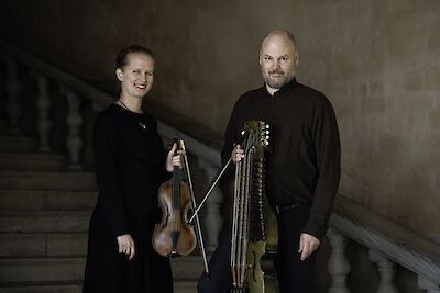 Das Bild zeigt zwei Musiker in schwarzer Kleidung, die in einem historischen Treppenhaus mit Steinwänden stehen: eine lächelnde Frau mit einer Violine und ein Mann mit Vollbart, der eine Nyckelharpa hält. Im Hintergrund sind die steinernen Stufen und das Geländer einer klassischen Architektur zu sehen.