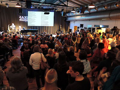 Das Bild zeigt eine gut besuchte Musikveranstaltung in einem industriell gestalteten Raum mit sichtbaren Lüftungsrohren und Bühnenbeleuchtung. Zahlreiche Besucher haben sich versammelt, während im Hintergrund Musiker auf einer kleinen Bühne auftreten und Liedtexte auf eine große Leinwand projiziert werden.