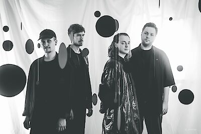 Ein schwarz-weißes Pressefoto zeigt vier junge Personen in dunkler Kleidung, die vor einem weißen Vorhang mit aufgedruckten schwarzen Kreisen posieren. Das Bild wirkt wie ein professionelles Bandfoto, bei dem die Personen ernst und direkt in die Kamera blicken.