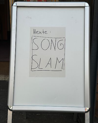 Das Bild zeigt einen weißen Werbeaufsteller mit einem handgeschriebenen Zettel, auf dem "Heute: SONG SLAM" steht. Der Aufsteller befindet sich vor einem Gebäude und wirbt für eine Song Slam Veranstaltung.