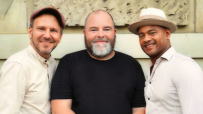 Drei Männer stehen nebeneinander. Sie lächeln in die Kamera. Zwei Männer tragen Hüte. Die Männer stehen vor einer hellen Wand. Sie sehen gut gelaunt aus.