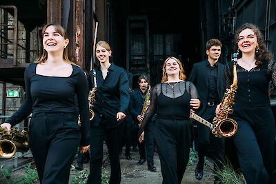 Eine Gruppe junger Musiker läuft durch ein altes Gebäude. Sie tragen schwarze Kleidung. Sie lachen und spielen Saxophone und andere Blasinstrumente. Das Gebäude ist alt und rostig. Die Musiker sind fröhlich.